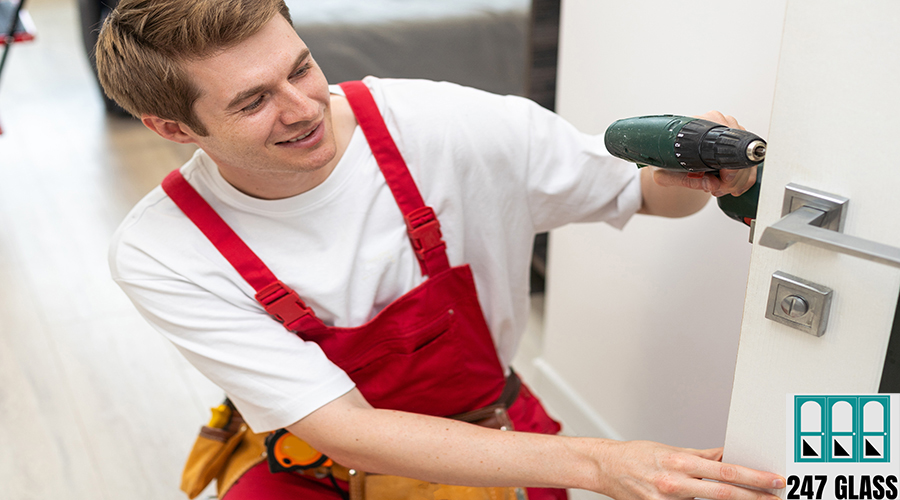 Installation of a lock on the front wooden entrance door. Portrait of young locksmith workman in blue uniform installing door knob. Professional repair service. Maintenance Concept door installation