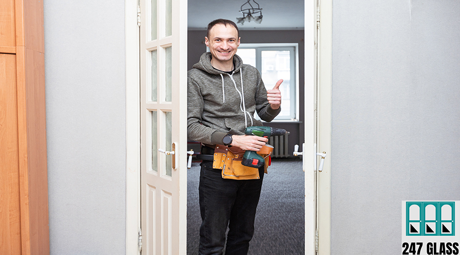 Maintenance Concept. Focused serious young locksmith with tattoo on hand wearing blue uniform standing on one knee in doorway installing doorknob handle, repairing wooden front door entrance door installation
